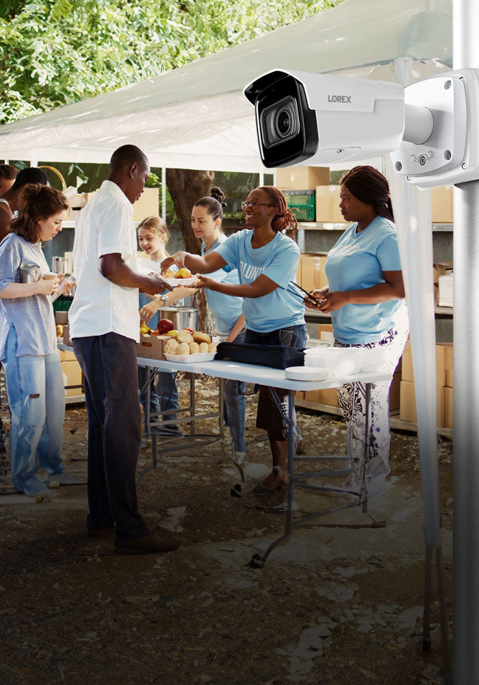 Lorex bullet security camera mounted on a pole, overlooking a group of volunteers serving food at an outdoor community event supporting Lorex for Business Not-For-Profit program