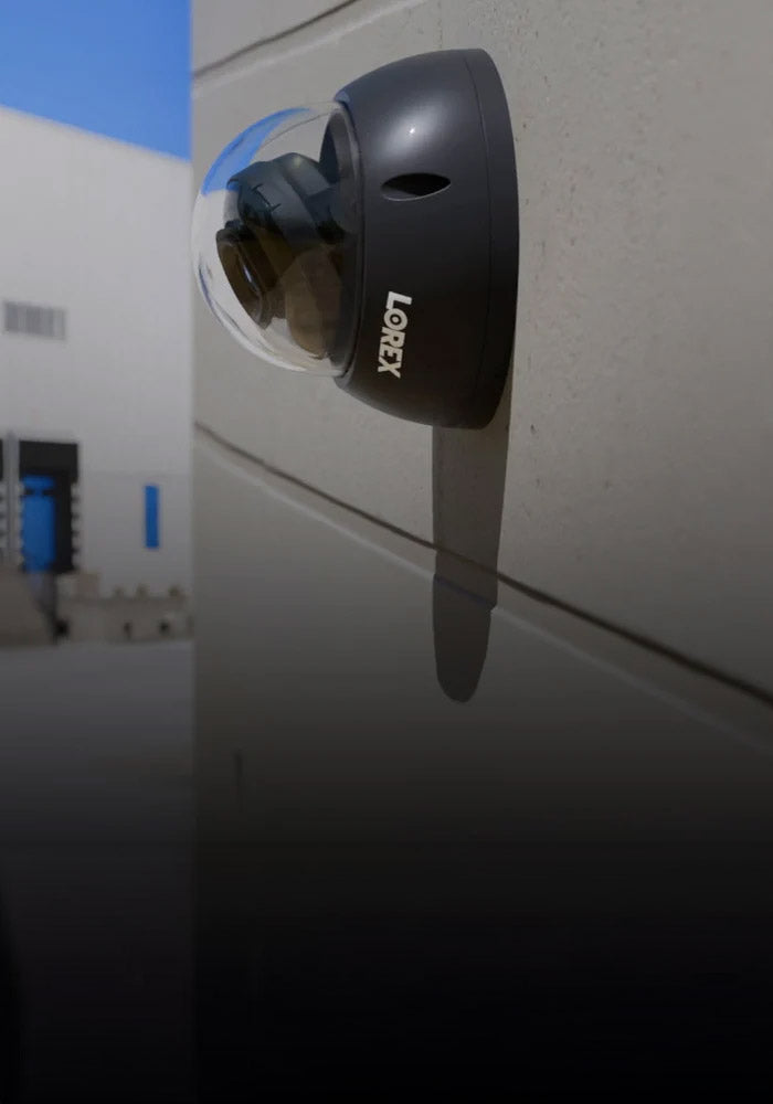 Lorex black dome security camera mounted on an exterior wall, overlooking a row of loading docks at an industrial warehouse under a clear blue sky.