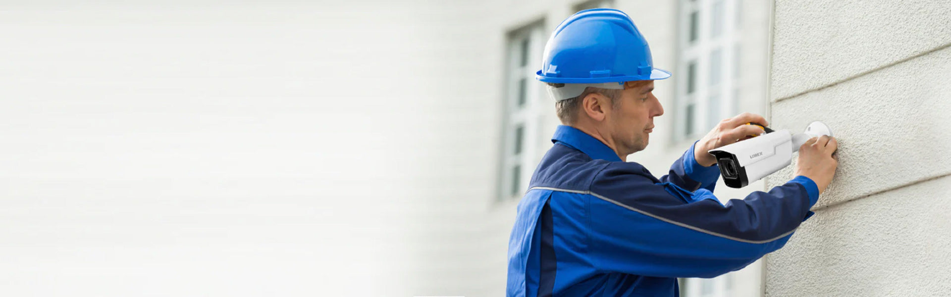 Professional installer in a blue uniform and hard hat mounting a Lorex security camera on an exterior wall, promoting the Lorex Installer Network for security professionals.